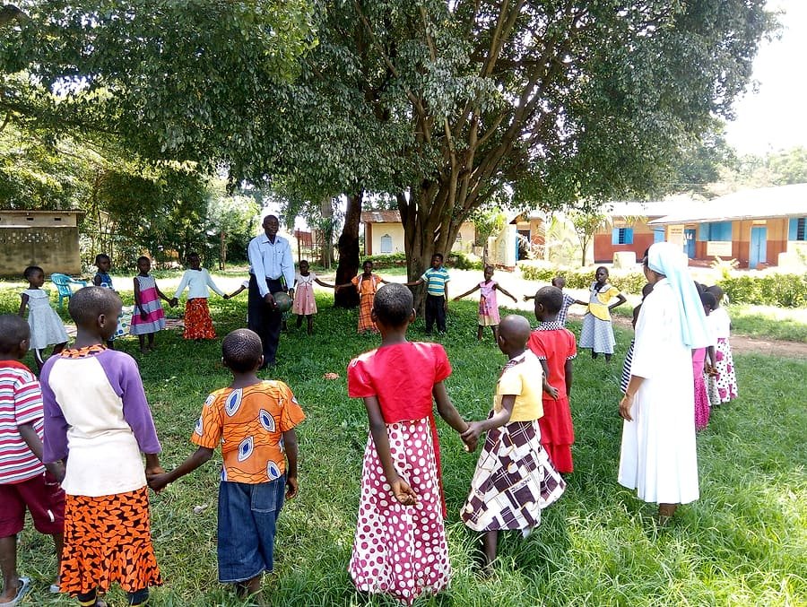 Children and community members standing in a wide circle under trees during a Kiyunga Social Service Centre community outreach session.