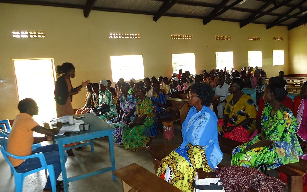 A facilitator addressing a full community hall of women and young people during a Kiyunga Social Service Centre community meeting.