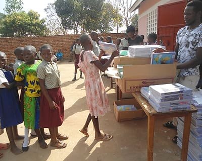 A KSSC team member handing exercise books to pupils queuing beside a table stacked with scholastic materials.