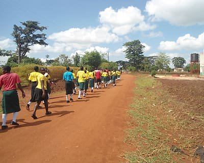 School pupils in uniform walking in procession along a village road during a community awareness march.